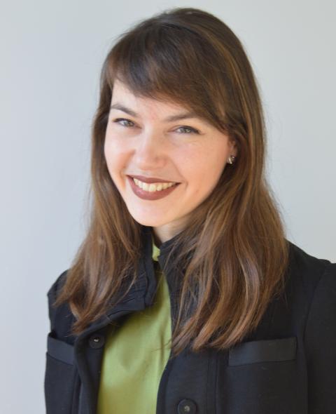 Hannah Levinson A feminine-presenting person with brown hair that falls just past their shoulders smiles into the camera. They have side bangs parted on the left and a light skin tone. They are wearing a green blouse with a thick black blazer on top, and the blazer has two pockets on either side.