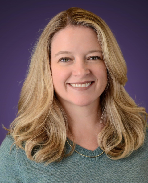 Blonde-haired, blue-eyed woman wearing a green shirt in front of a purple backdrop