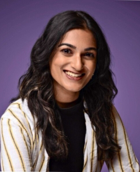 Brunette woman wearing a black t-shirt and white dress shirt with yellow vertical strips in front of a purple backdrop