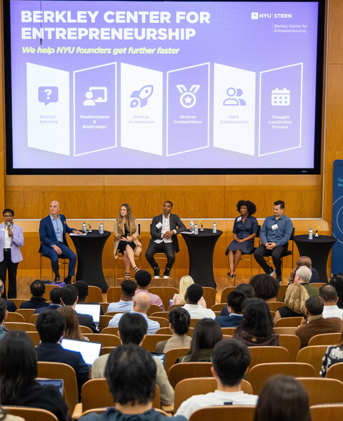 Audience in a large auditorium listening to a panel on a stage.
