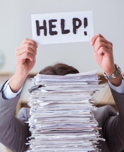 Man holds up "Help!" sign while hidden behind pile of papers