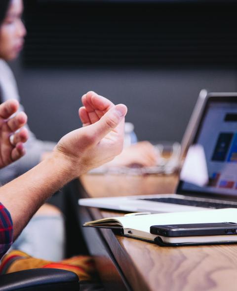 Abstract photo of hands gesturing while speaking at a table showing a laptop, notebook, and phone
