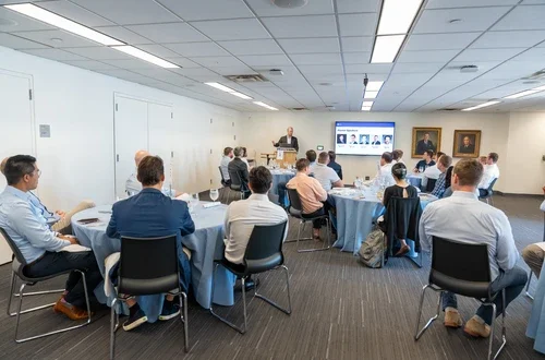 A group of students seated at round tables, attentively listening to a speaker at the front of the classroom
