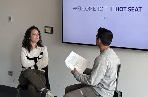 A feminine-presenting person with fair skin and curly, shoulder-length hair sits with her arms crossed across from a masculine-presenting person with medium-tan skin and dark hair, as he olds a piece of paper in his hands and gestures while talking. A screen behind these two reads, "Welcome to the hot seat."