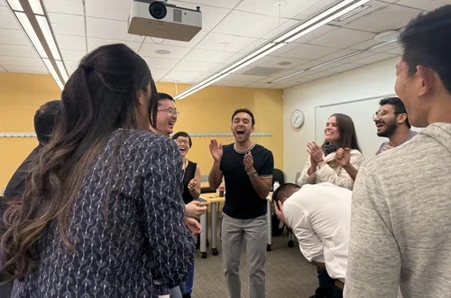 A group of seven students stands in a circle clapping and laughing after a game of rock paper scissors.