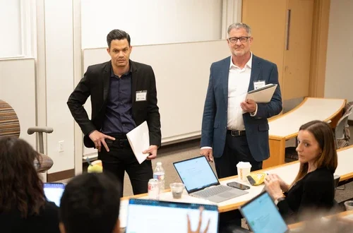 Two masculine presenting people with lighter skin stand in blazers as they hold a pile of papers. One stands with his right hand on his hip. They listen to a small group of seated people in a lecture-style classroom.  