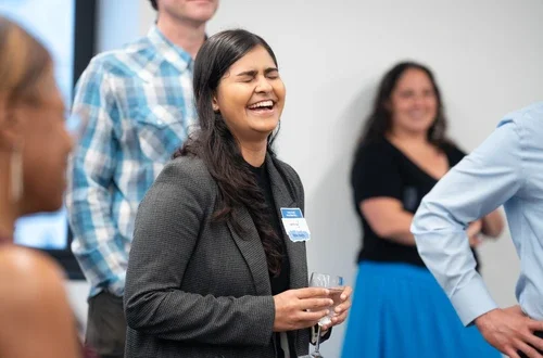 A feminine-presenting person with long, black, curled hair laughs so had their eyes close. They hold a cup in their hands and are surrounded by others in a sparse, standing crowd.