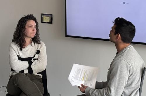 A feminine-presenting person with light skin and dark, curly hair crosses her arms over her chest as she stares with muted animosity at her partner, seated across from her with dark skin, short black hair, and a thick black mustache. 