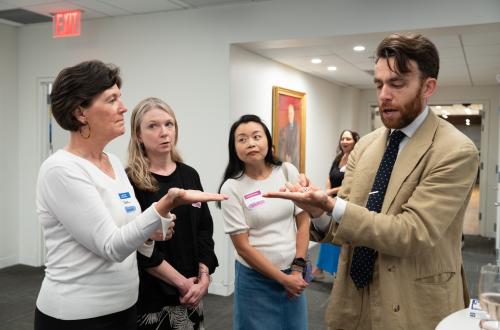 A masculine-presenting magician with light skin dressed in a khaki suit holds out his hand with palm turned up to the sky. A group of feminine-presenting people with light skin gather round interested. One of the observers holds her palm turned up to the sky as she looks at the magician.