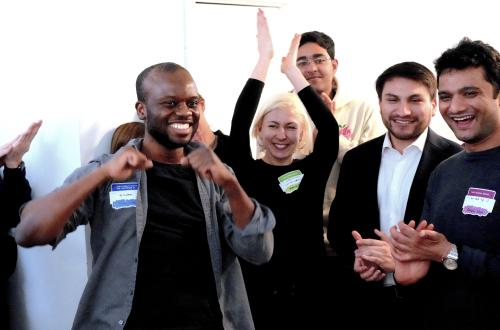 A masculine-presenting person smiles at the camera with their thumbs pointing at their chest. They are surrounded by a group of fellow students, who smile and cheer him on.