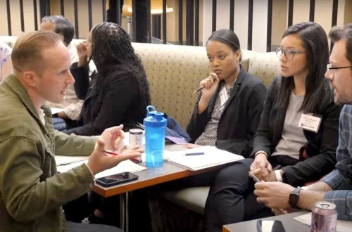 A group of four people huddle, seated around a table with paper and pens in hands. They are planning.