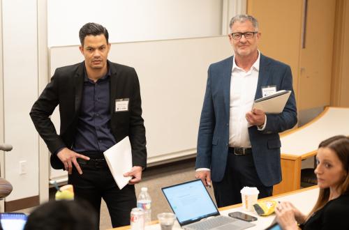 Two masculine presenting people with lighter skin stand in blazers as they hold a pile of papers. One stands with his right hand on his hip. They listen to a small group of seated people in a lecture-style classroom.  
