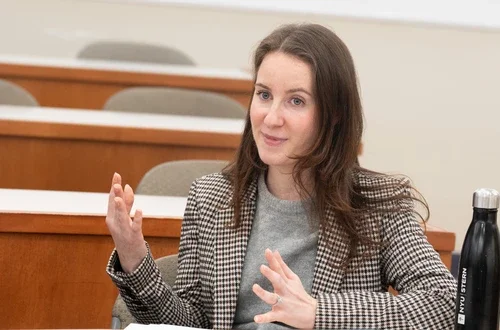 A feminine-presenting person with medium length hair sits at a desk and speaks with her hands moving in front her body as she does.