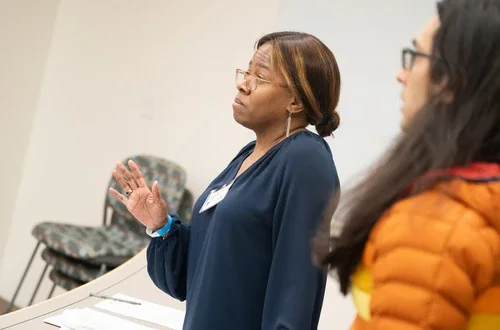 A feminine-presenting person stands with their eyebrows raised and hand raised in defense. 