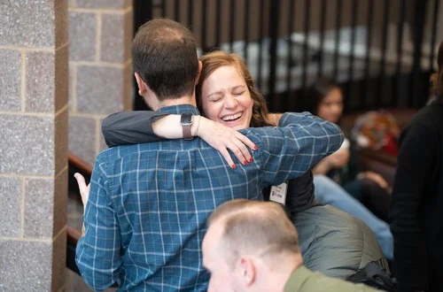 One feminine and one masculine presenting person hug each other, smiling.
