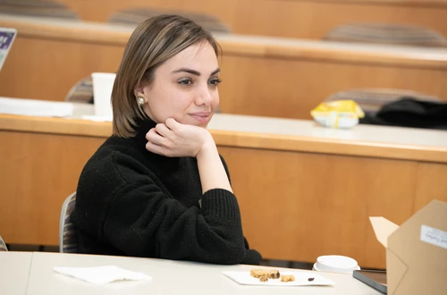 A feminine-presenting person with pale skin and highlighted hair sits in a classroom with her head resting in her palm, engaged. 