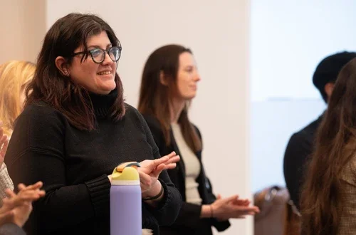 A feminine presenting person with light skin and dark, shoulder-length hair wearing glasses smiles and claps in a crowd of standing people.