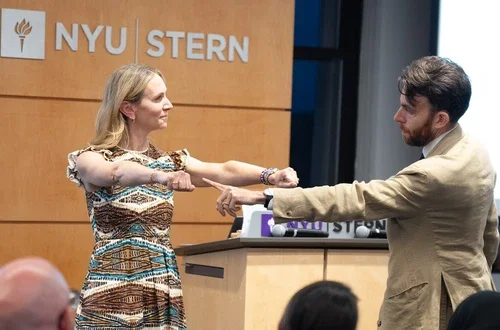 A masculine presenting magician points his fingers at the right fist of a feminine presenting person, who stands with her arms outstretched in front of her.