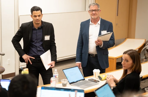 Two masculine presenting people with lighter skin stand in blazers as they hold a pile of papers. One stands with his right hand on his hip. They listen to a small group of seated people in a lecture-style classroom.  