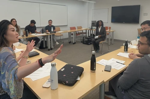 A group of twelve students addresses a feminine-presenting actor seated in the middle of the room. 