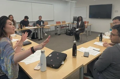 A group of twelve students addresses a feminine-presenting actor seated in the middle of the room. 