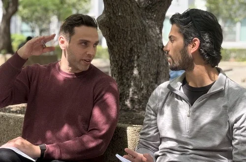 Two masculine-presenting students sit outside under a tree in Sasaki Garden. They have notebooks in their hands and they give each other eye contact, as well as feedback.