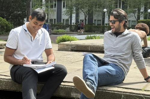Two masculine-presenting people with tan skin and black hair sit outisde smiling, as the figure on the left writes in his notebook. 