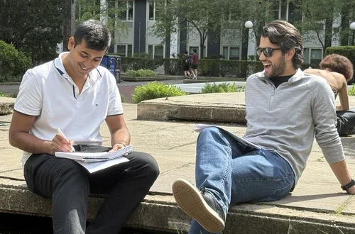 Two masculine-presenting people with tan skin and black hair sit outisde smiling, as the figure on the left writes in his notebook. 