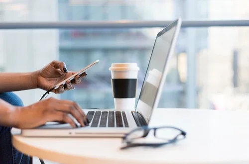 Person's hands on a phone with a laptop, cup of coffee, and pair of glasses in the frame