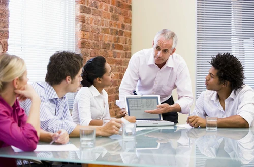 four people sitting at a glass table looking at a fifth person who is explaining something from a laptop