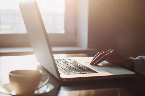 hand on touchpad of a laptop, next to a mug, in a room with sunshine beaming in