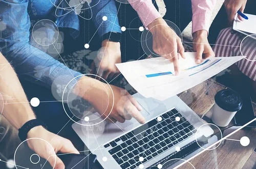 group of people's arms at a table, with one person pointing at a laptop and another person holding and pointing at a sheet of paper