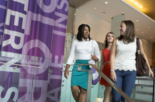 three women in business casual attire looking at each other as they descend a staircase