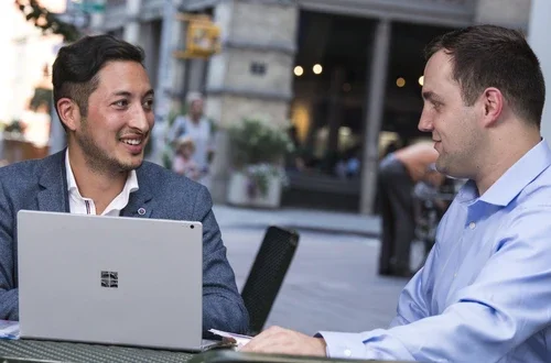 two young men in business clothing sitting at a table with a laptop