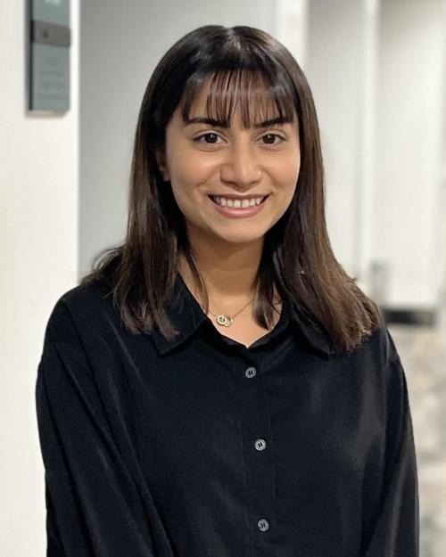 A feminine-presenting person with medium-dark skin tone, and short, straight black hair, and bangs smiles into the camera. She is wearing a black silk collared shirt and standing in the middle of a long empty hallway.
