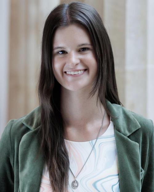 A feminine-presenting person with long brown hair parted down the middle smiles into the camera. They are wearing a velvet green blazer and white crewneck shirt with thin wavy lines of various colors dancing along the bodice. They are wearing a long silver necklace, and they are standing in front of a row of large Greek columns. 