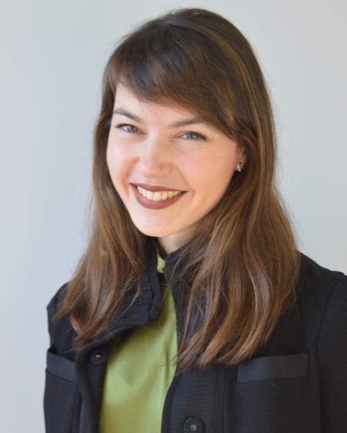 A feminine-presenting person with brown hair that falls just past their shoulders smiles into the camera. They have side bangs parted on the left and a light skin tone. They are wearing a green blouse with a thick black blazer on top, and the blazer has two pockets on either side.