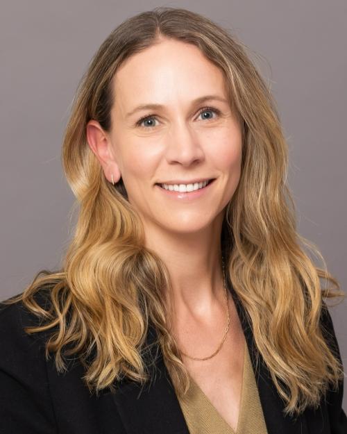 A feminine-presenting person with light skin tone and wavy blond hair that falls past her shoulders smiles into the camera. She is wearing a black blazer with sleeves rolled over her elbows, and her gold blouse has a line of small buttons running down the middle. 