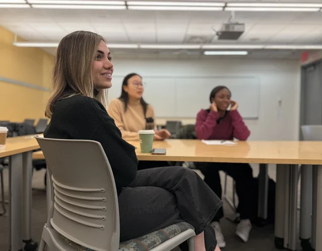 Three feminine-presenting students sit in a half-moon and smile in a classrom.