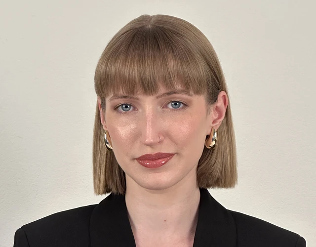 A feminine-presenting person with light skin and hair smiles softly into the camera. She's wearing an oversized blazer, and her hair is in a bob.