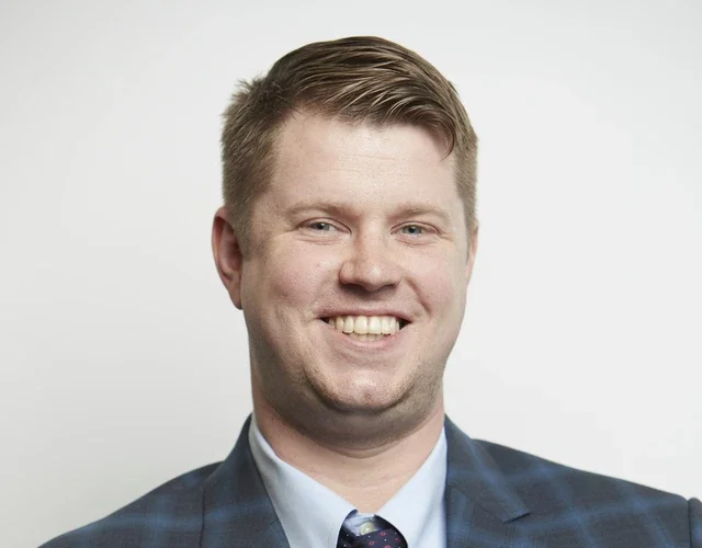 A masculine-presenting person with light skin and hair wears a plaid suit and tie as he smiles directly into the camera.