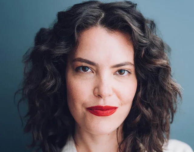 A feminine-presenting person with light skin and dark curly hair smiles softly into the camera. She's wearing a white short-sleeved collared shirt and red lipstick. 