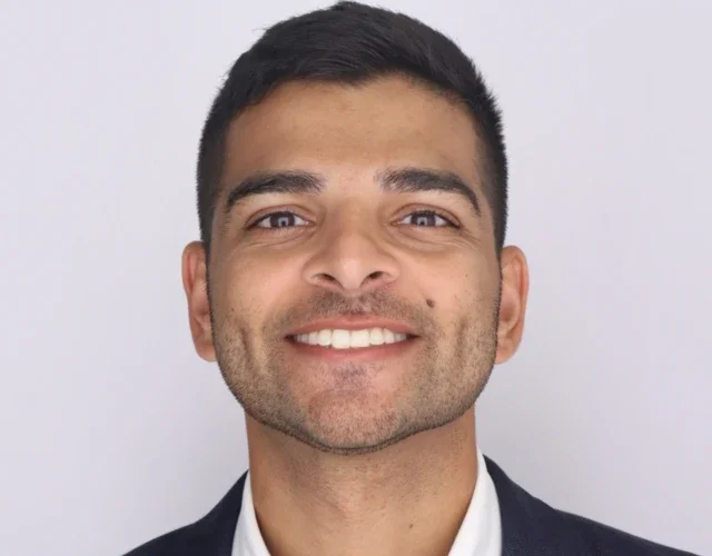 A masculine-presenting person with medium tan skin and dark hair wears a dark blue blazer and white collared shirt as he smiles directly into the camera.