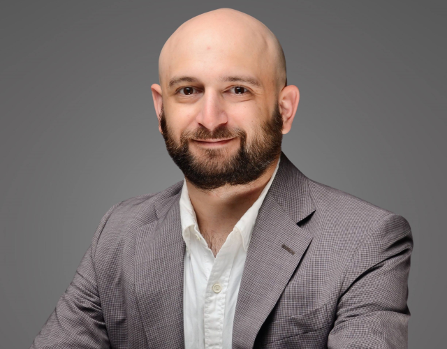 A bald person with a dark goatee crosses his hands over his knees as he sits in a gray suit. His name is Micah.