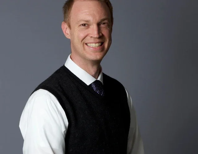 A masculine-presenting person with light skin smiles directly into the camera. He wears a crisp white button-down with a black sweater-vest on top.