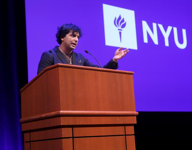 Filmmaker M. Night Shyamalan speaking at a podium in front of NYU logo background