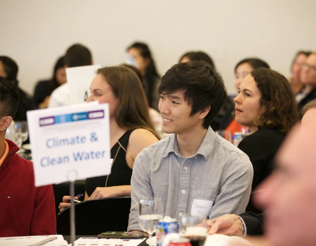 Young man in focus sitting at a table with a sign reading Climate & Clean Water. 