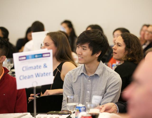 Young man in focus sitting at a table with a sign reading Climate & Clean Water. 
