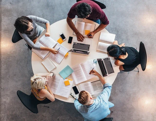 aerial view of five people sitting at a round table, with laptops, books, and notes strewn about