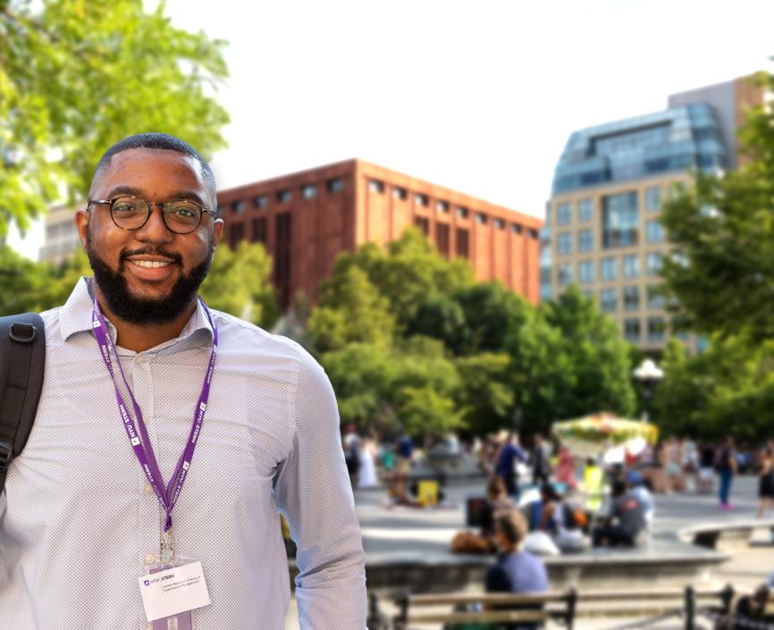 Male student with backpack, light color shirt and a New York University ID hanging from his neck in front of blurred background image of Stern School of business seen from the fountain at Washington Square Park
